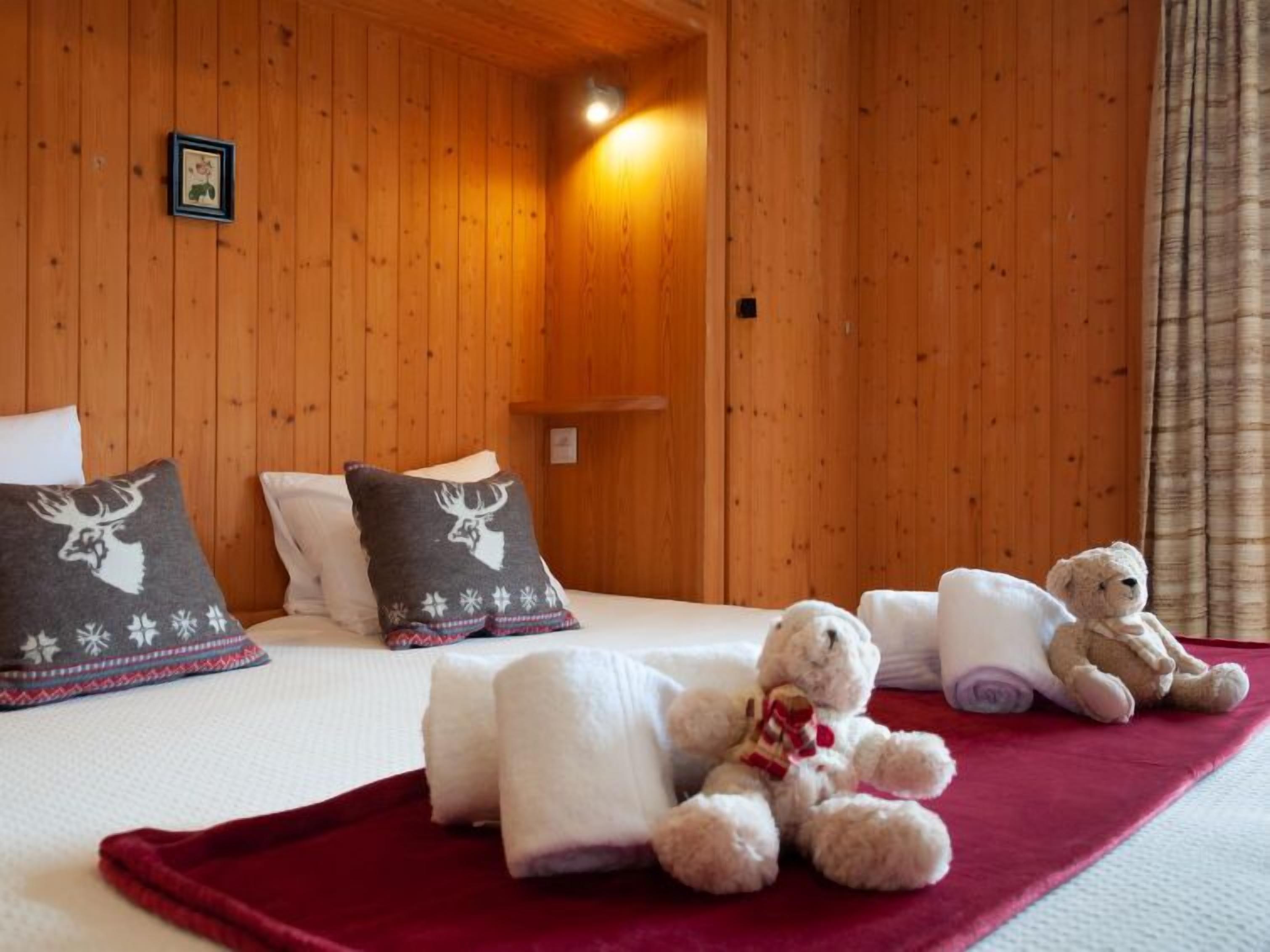 Ein gemütliches Zimmer mit Holzwänden, einem Bett mit weißer Bettwäsche, zwei Kopfkissen mit Hirschmotiven und zwei Teddybären auf dem Bett.