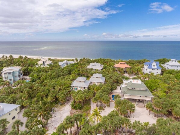 Ett flygbildsbild av ett bostadsområde vid kusten, med hus omgivna av tät vegetation och en strand i bakgrunden.