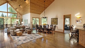 A spacious living area with a high ceiling, wooden flooring, and large windows offering views of the outdoors.