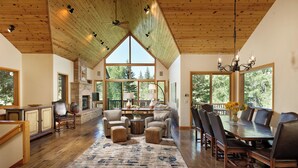 A spacious living room with a fireplace, wooden flooring, and large windows offering a view of trees.