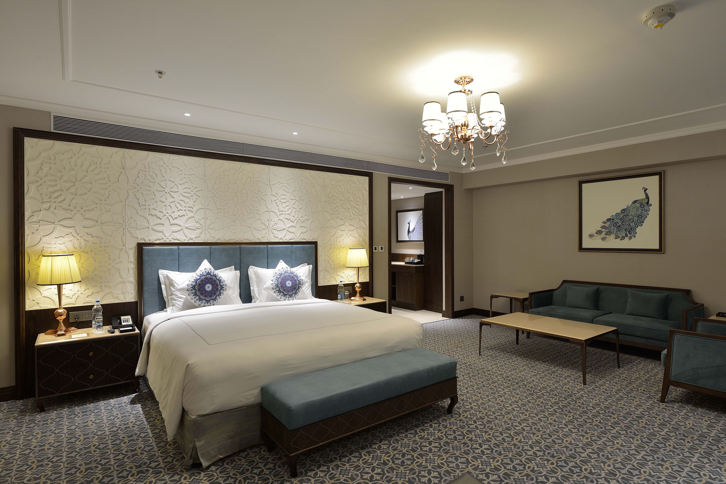 A hotel room with a large bed, two bedside tables with lamps, a bench, a sofa, a TV, and a chandelier.