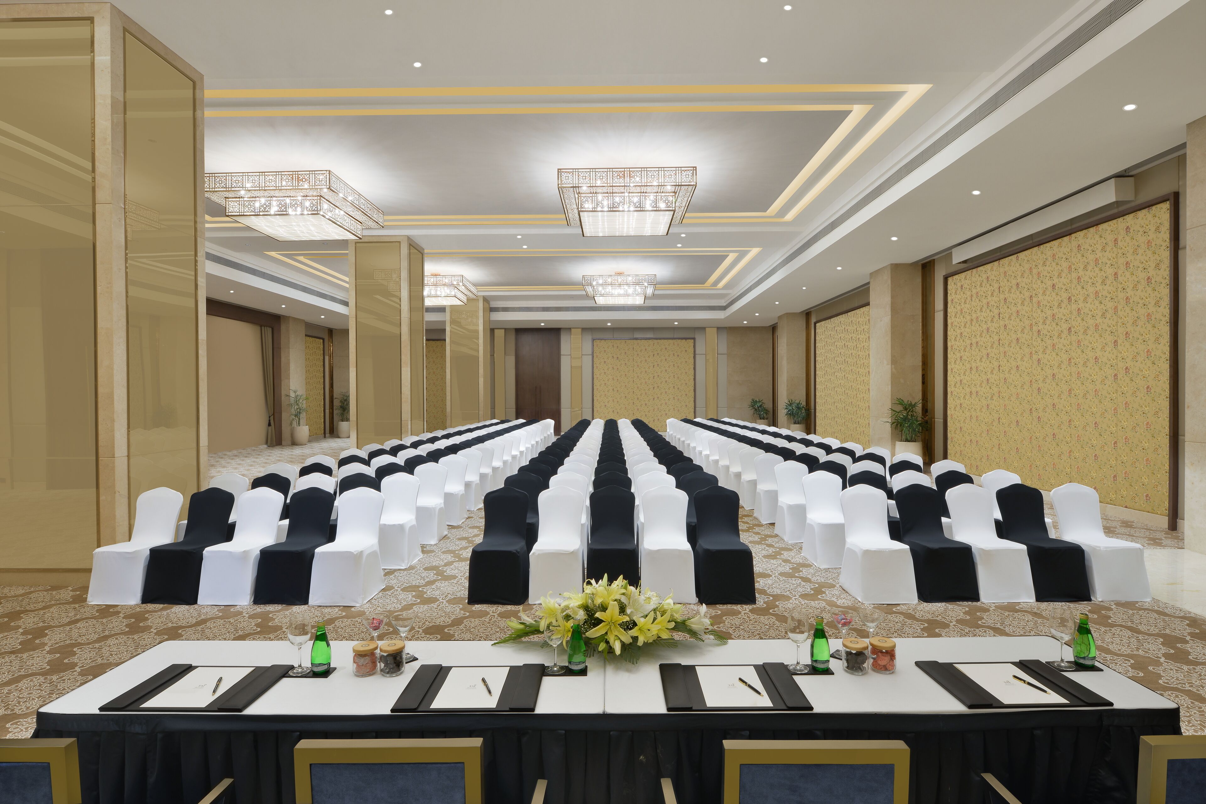 Une salle de conférence avec des rangées de chaises blanches et noires, une longue table ornée de plaques nominatives et un lustre.