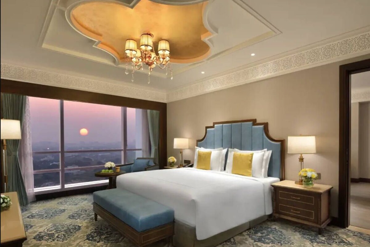 A hotel room with a large bed, a view of the sunset through the window, a chandelier, and a patterned carpet.
