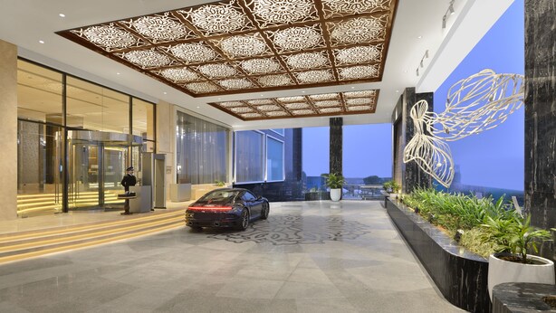 A modern hotel lobby with a car parked in the center, a security guard, and a decorative ceiling.