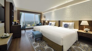 A hotel room with two beds, a desk, a chair, a TV, and a large window with a view.