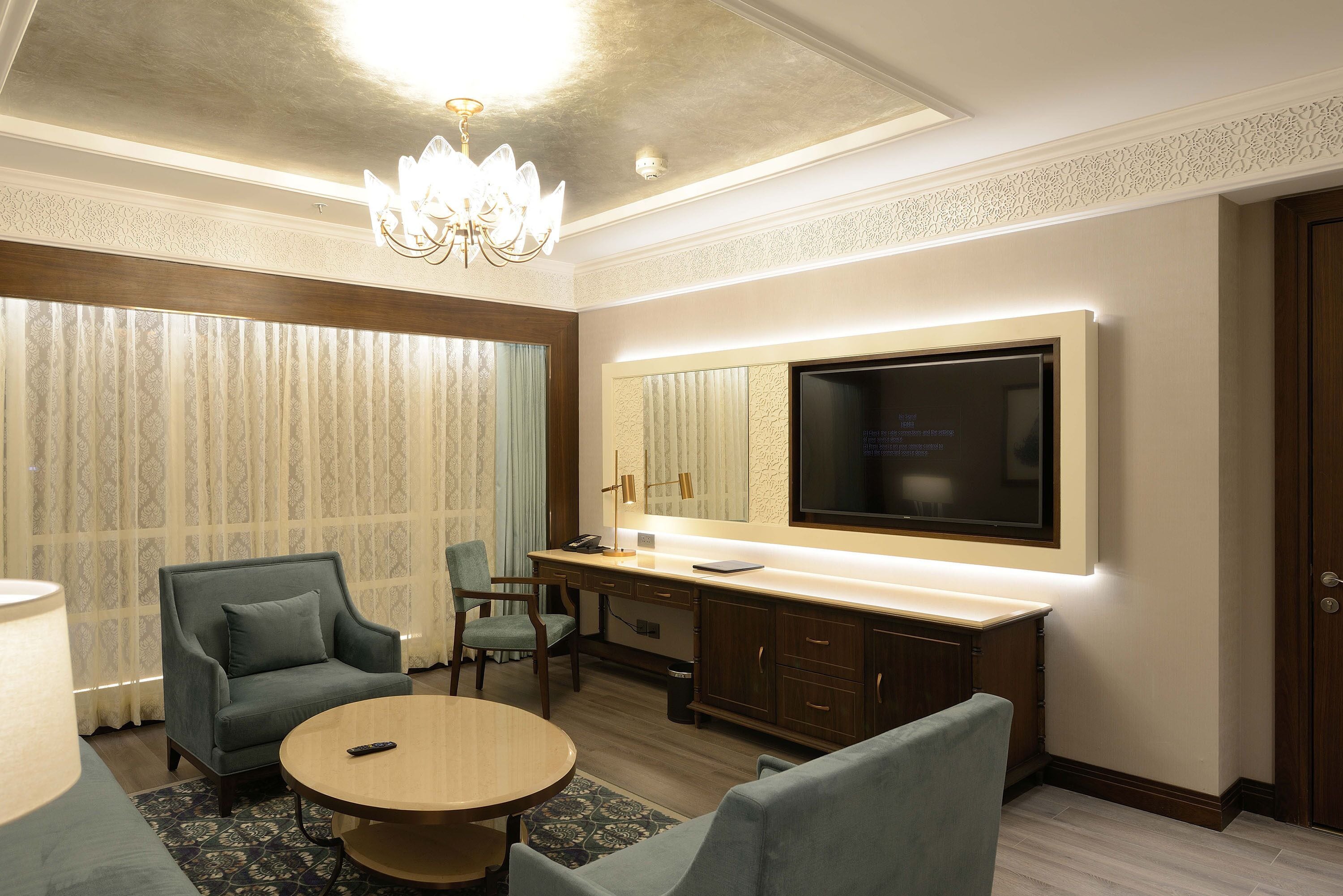A modern hotel room with a large flat-screen TV, a desk with a lamp, two armchairs, and a round table.