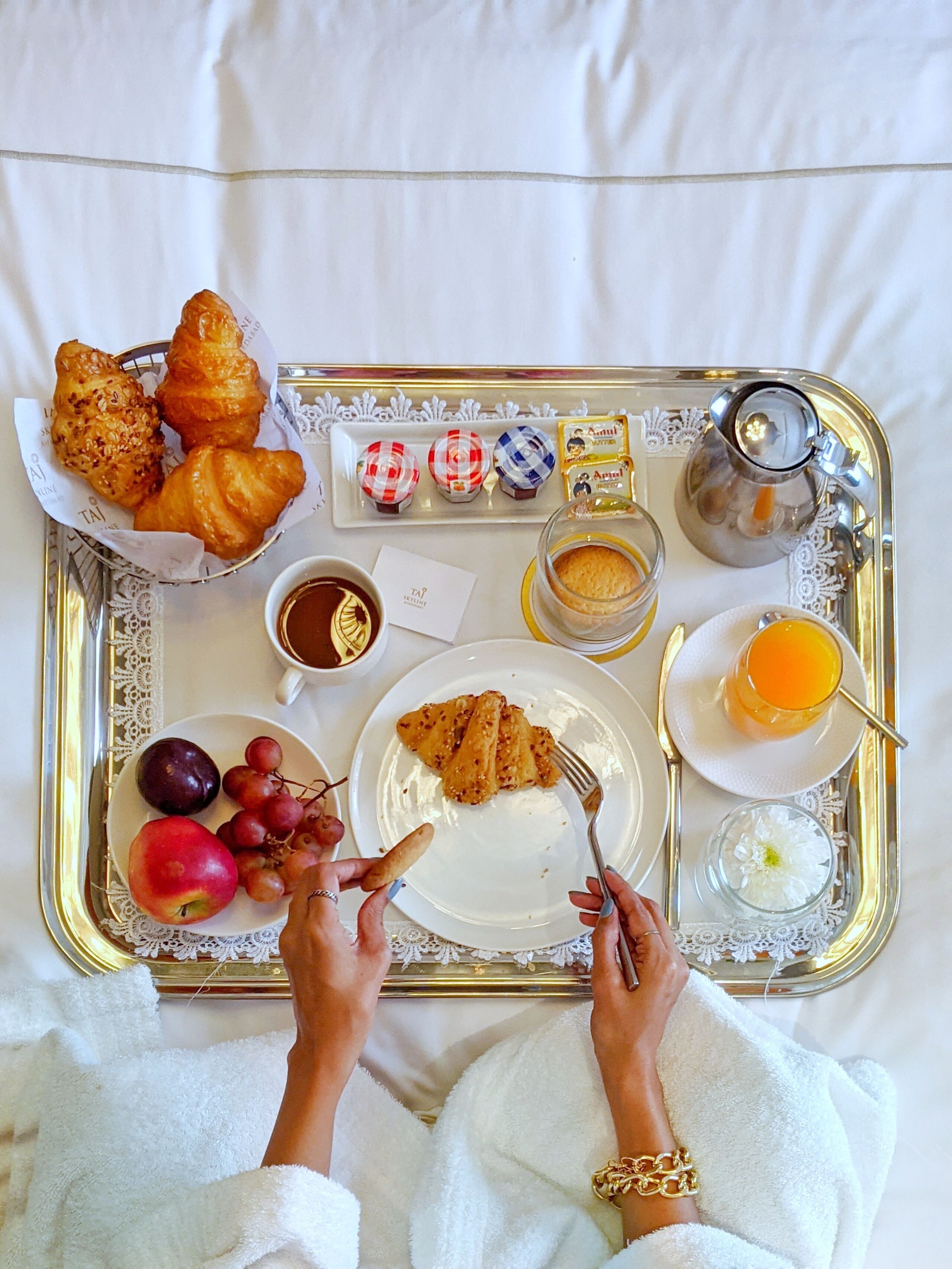 A breakfast tray with croissants, pastries, fruit, and beverages on a white bedspread.