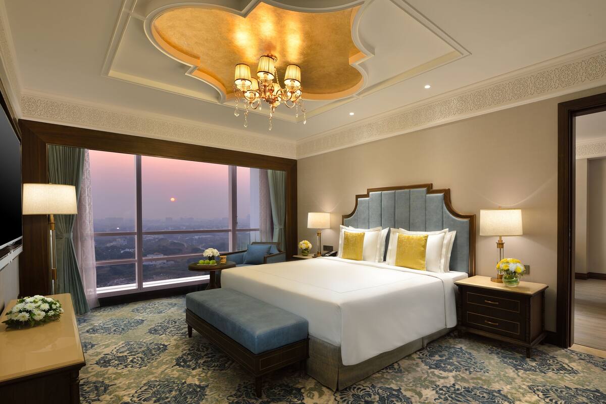Een hotelkamer met een groot bed, uitzicht op de stad, een kroonluchter en een bureau met een lamp.