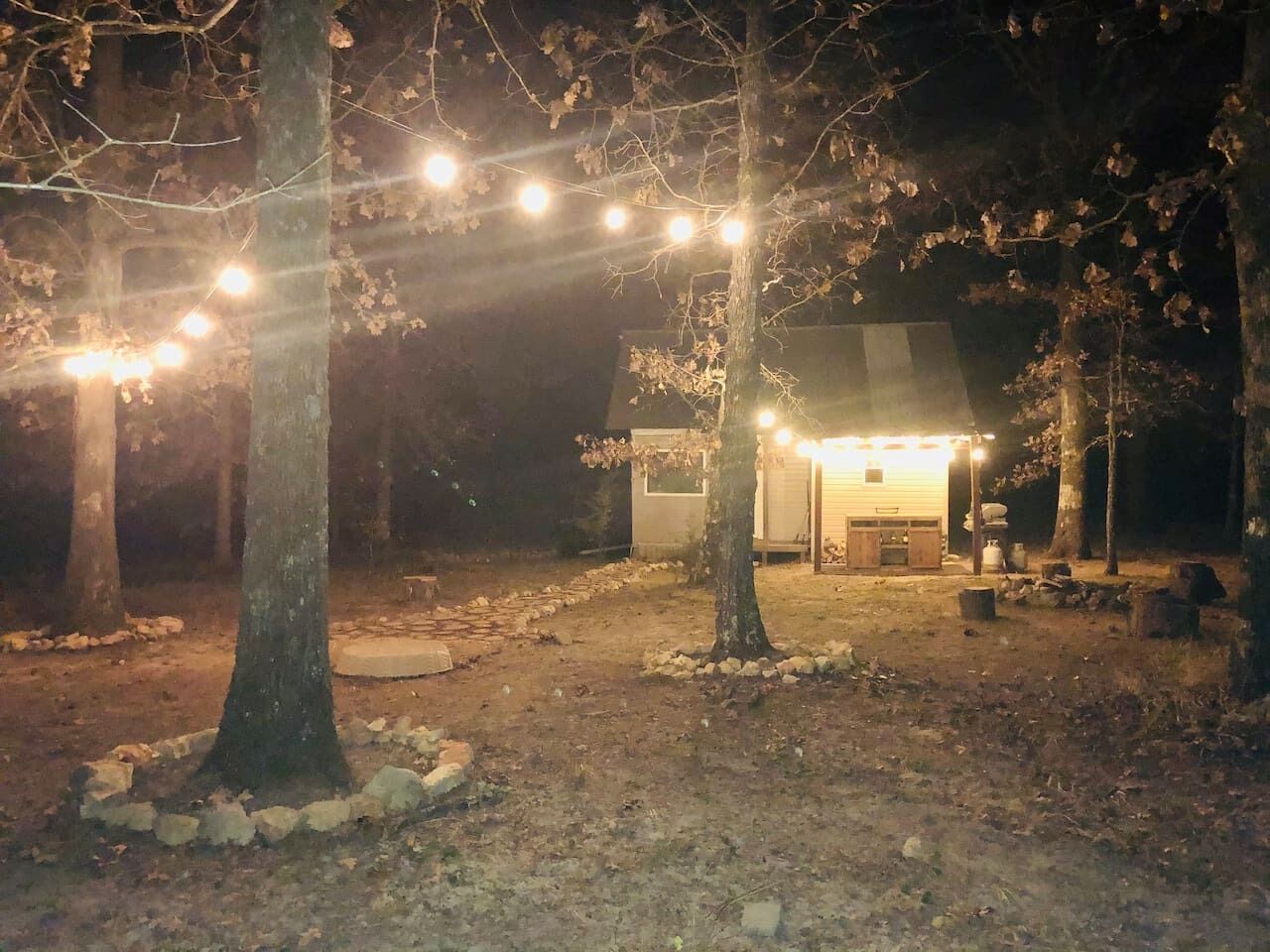 A rustic cabin with string lights, surrounded by trees, and a small outdoor seating area.