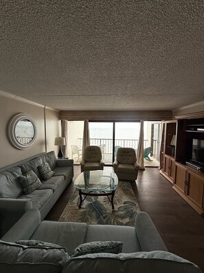 Una sala de estar con un sofá, sillones, una mesa baja de vidrio, una televisión y vista al mar a través de ventanas amplias.