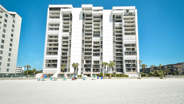 Un hotel moderno de varios pisos con balcones, situado cerca de una playa con sombrillas y palmeras.