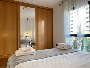 Ein Hotelzimmer mit großem Spiegel, einem Bett mit ordentlich gefalteten Handtüchern, einem Nachttisch und Blick auf ein Gebäude durch leichte Vorhänge.