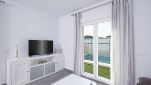 Ett modernt vardagsrum med en platt-TV, ett vitt soffbord och en skjutdörr i glas som leder ut till en pool.