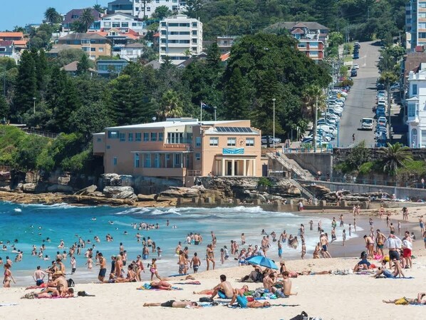 A beach with people sunbathing and swimming, a building with a blue sign, and a road with parked cars.