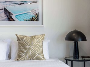 A hotel room with a bed, a pillow, a lamp, a framed picture of a pool, and a small table.