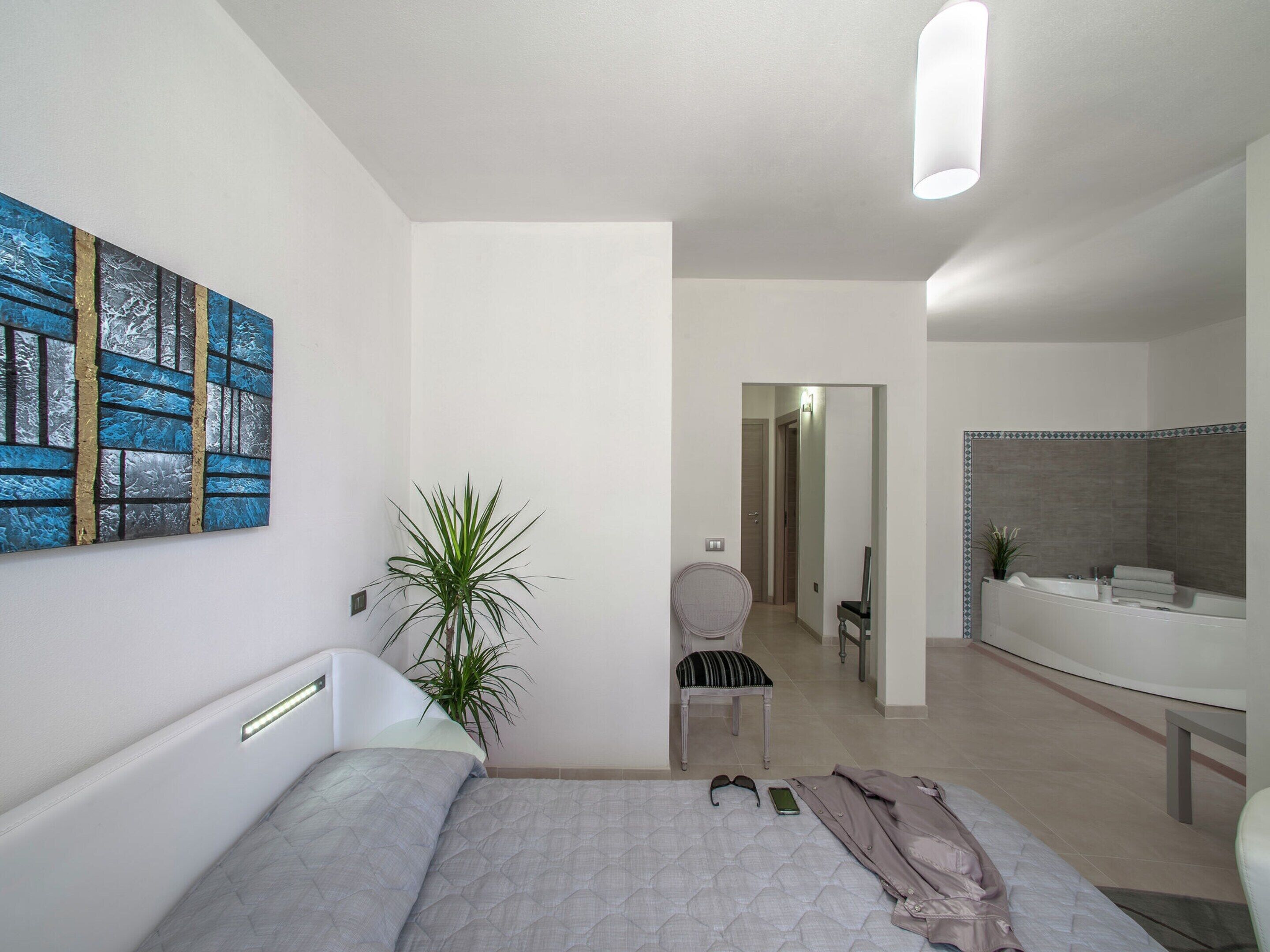 A modern bedroom with a bed, a bedside table, a chair, a plant, and a bathroom with a bathtub and mirror.
