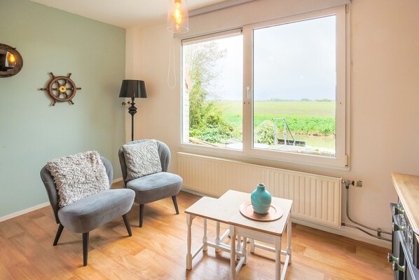 A room with two grey armchairs, a small white table, a lamp, and a view of a green field through a large window.