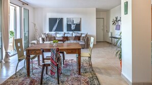 A modern living room with a wooden dining table, chairs, a sofa, and a coffee table. There are two large windows with curtains, a radiator, and a wall-mounted light fixture. - Beautiful Stay in Tavullia (Tavullia)