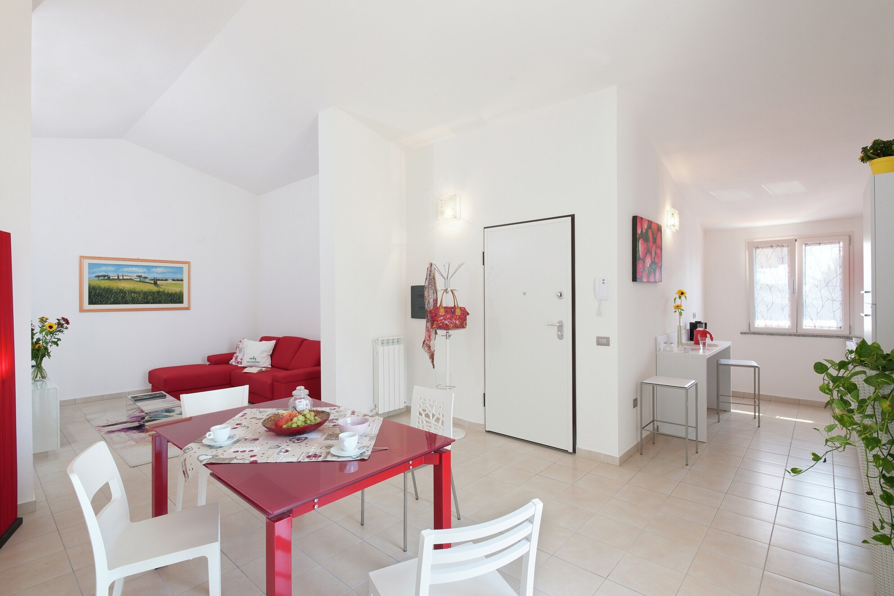 Un vaste espace de vie comprenant un coin repas avec une table rouge, des chaises blanches, un canapé rouge et un coin cuisine doté d’un comptoir blanc et de deux tabourets de bar.