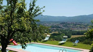 A rectangular swimming pool with a red slide, surrounded by grass and trees, with a view of a town and mountains in the background.