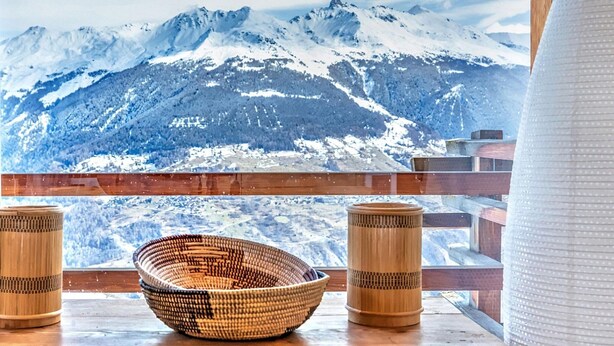 A wooden table with a woven bowl and two bamboo candle holders, overlooking a snowy mountain range. - Stylish Chalet With Sauna in Heremence (Heremence)