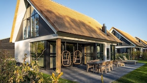 A modern house with a large glass window, a thatched roof, and a patio with outdoor furniture. - Luxury Villa With Sauna Near the Beach (De Cocksdorp)