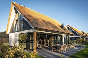 Ein modernes Haus mit einem großen Glasfenster, einem Strohdach und einer Terrasse mit Gartenmöbeln.