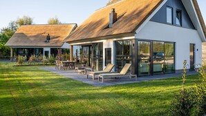 A modern house with a thatched roof, large glass windows, and a well-maintained lawn. - Luxury Villa With Sauna Near the Beach (De Cocksdorp)