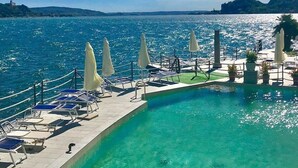 Ein Poolbereich mit Liegestühlen und Sonnenschirmen, mit Blick auf ein Gewässer, im Hintergrund sanfte Hügel.