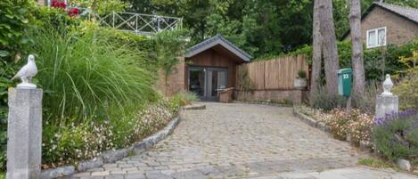 A cobblestone pathway leading to a wooden building with a sloped roof, surrounded by lush greenery and a fence.