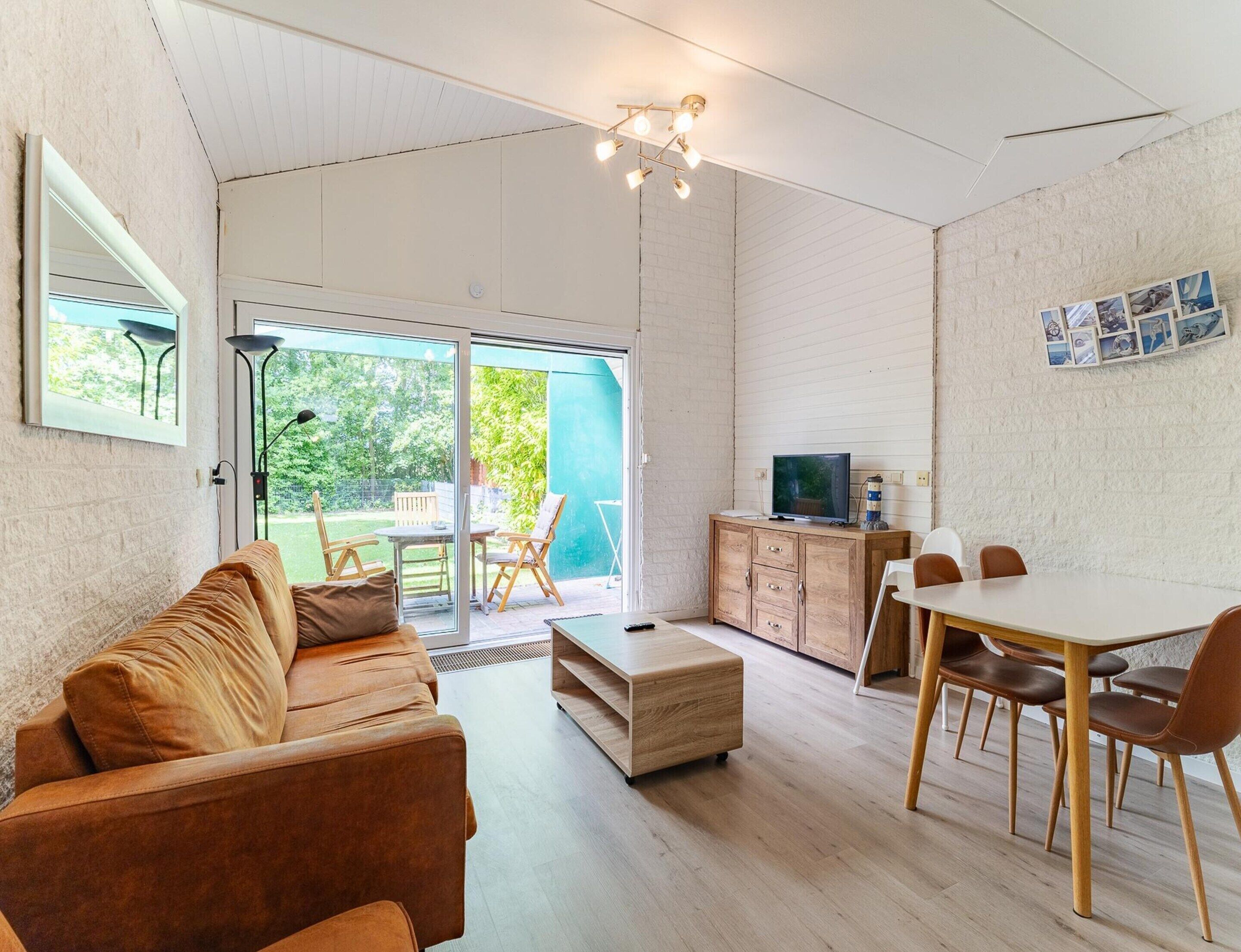 A modern living room with a sofa, a dining table with chairs, a television on a wooden cabinet, and a large sliding glass door leading to an outdoor area.