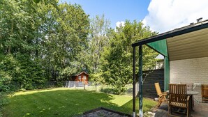 A covered patio area with outdoor seating and a view of a wooden cabin in a wooded area.