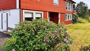 Et rødt toetagers hus med hvide døre og vinduer, en lille veranda og en velplejet have med buske og blomster.
