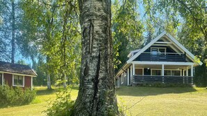 A modern two-story house with a large tree in the foreground, a grassy yard, and a small red building in the background. - 6 Person Holiday Home in Torsby (Torsby)