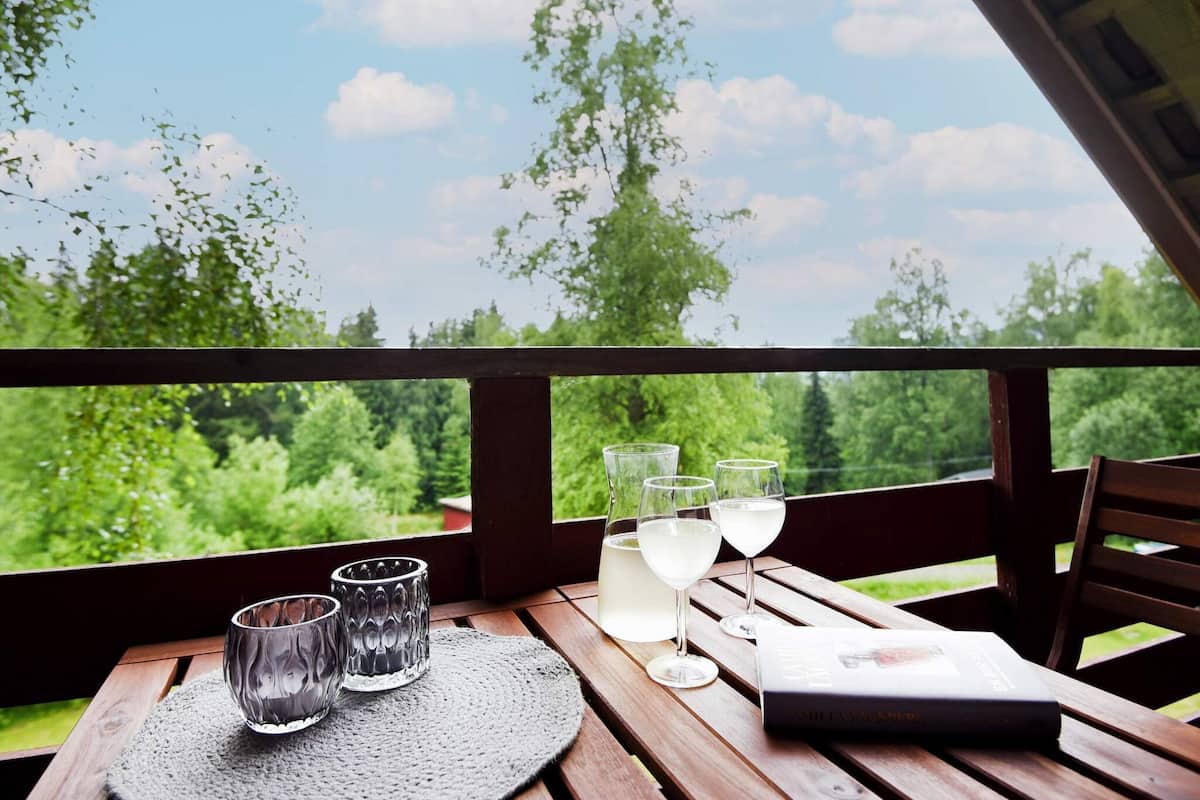 A wooden table with three glasses, a bottle, and a magazine on a balcony with a view of greenery and a clear sky.