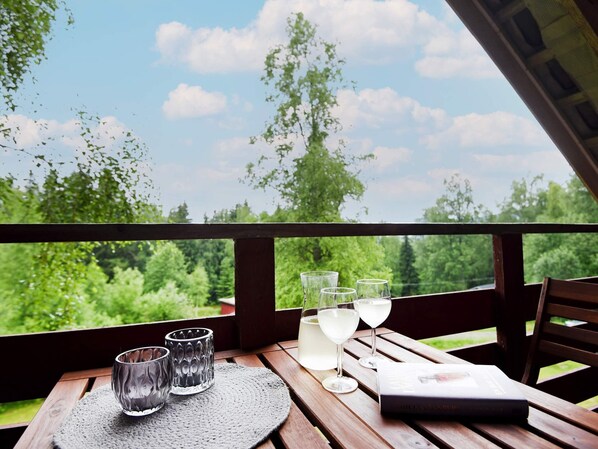 A wooden table with three glasses, a bottle, and a magazine on a balcony with a view of greenery and a clear sky. - 6 Person Holiday Home in Torsby (Torsby)