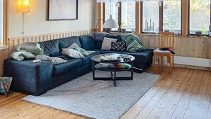 A cozy living room with a dark blue sectional sofa, a small round coffee table, and a white rug. There is a framed picture on the wall, a window with a view of trees, and a lamp on the side table. - 6 Person Holiday Home in Torsby (Torsby)