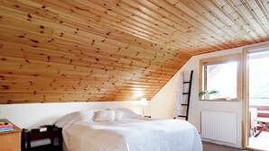A bedroom with a sloped wooden ceiling, a bed with white bedding, a bedside table with books, a lamp, and a window with a view. - 6 Person Holiday Home in Torsby (Torsby)