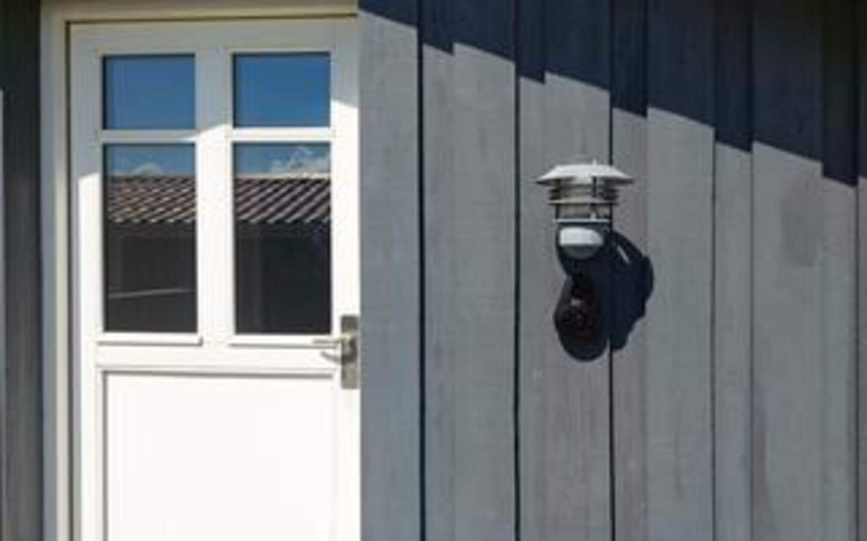 Une porte blanche avec une fenêtre, un luminaire fixé au mur et une façade d’immeuble de couleur sombre.