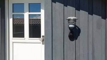 Une porte blanche avec une fenêtre, un luminaire fixé au mur et une façade d’immeuble de couleur sombre.