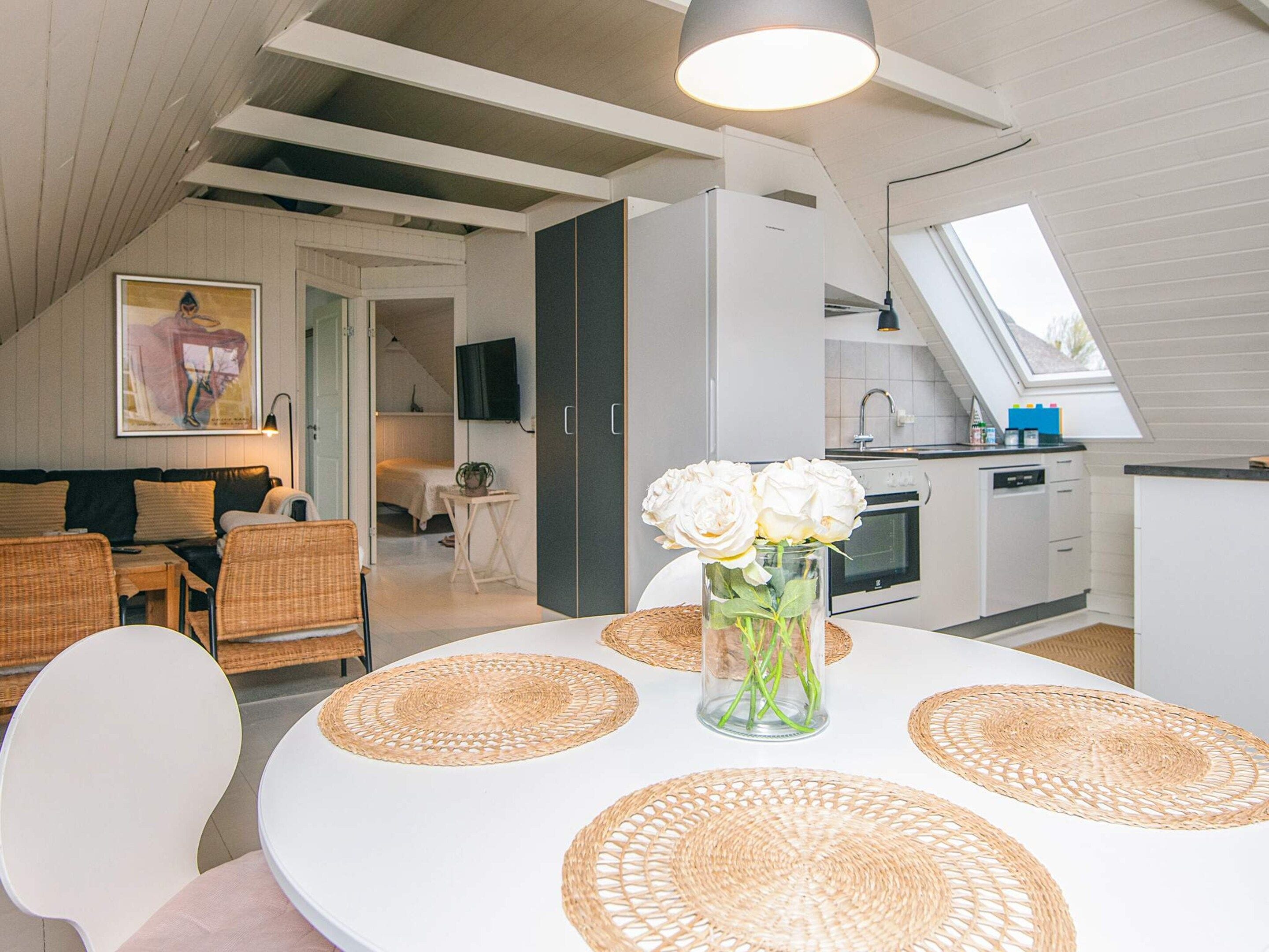 Una cocina moderna con tragaluz, un comedor con mesa redonda y individuales tejidos, y una sala de estar con sofá y sillones.