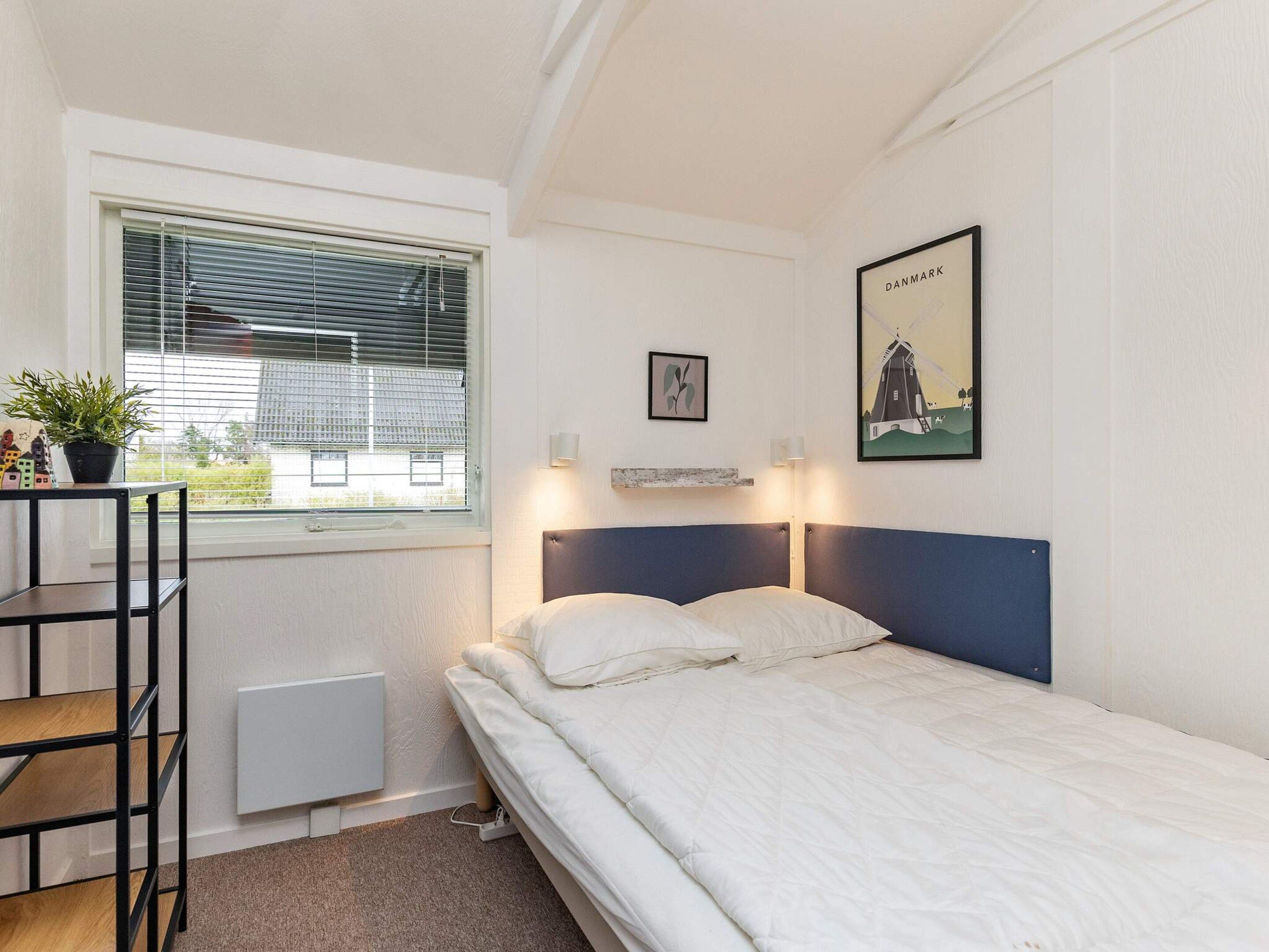 Uma cama de solteiro com cabeceira azul num quarto com paredes brancas, uma janela com persianas, um radiador, uma pequena mesa com uma planta e um quadro com uma imagem de um moinho.
