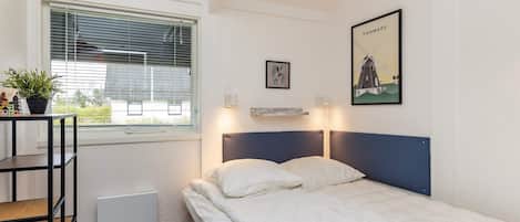 A single bed with a blue headboard in a room with white walls, a window with blinds, a radiator, a small table with a plant, and a framed picture of a windmill.