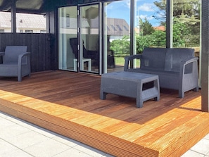 A covered outdoor seating area with wicker furniture, a wooden deck, and a glass door leading to a garden.