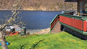 Una cabaña junto al lago con una terraza de madera y un ventanal de vidrio, con vistas a un lago tranquilo y un paisaje montañoso.