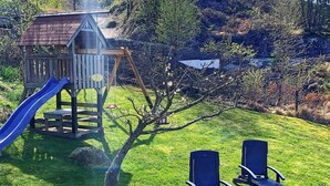 Una estructura de juego de madera con un tobogán y una casita pequeña, rodeada de césped y árboles.