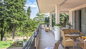 A covered outdoor area with wicker furniture, a large potted plant, and a view of a lush garden. - Grand Villa in Tavullia (Tavullia)
