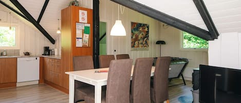 A dining area with a white table and brown chairs, a kitchen with wooden cabinets, and a television on a stand.