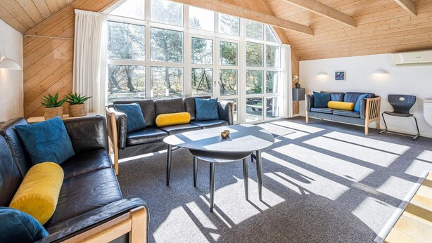 A spacious living area with a high ceiling and large windows, a black leather sofa, a small round table, and a blue couch with yellow cushions. - 10 Person Holiday Home in Norre Nebel (Norre Nebel)