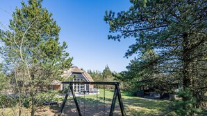 A house with a thatched roof, surrounded by a wooded area and a swing set. - 10 Person Holiday Home in Norre Nebel (Norre Nebel)
